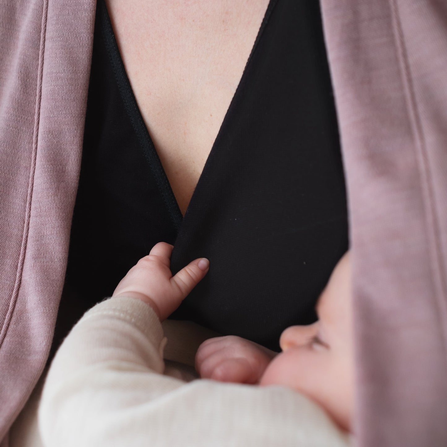 A close-up lifestyle shot of a baby wearing a cream long-sleeve outfit while nursing from a mother wearing a black V-neck top and a dusty rose cardigan.