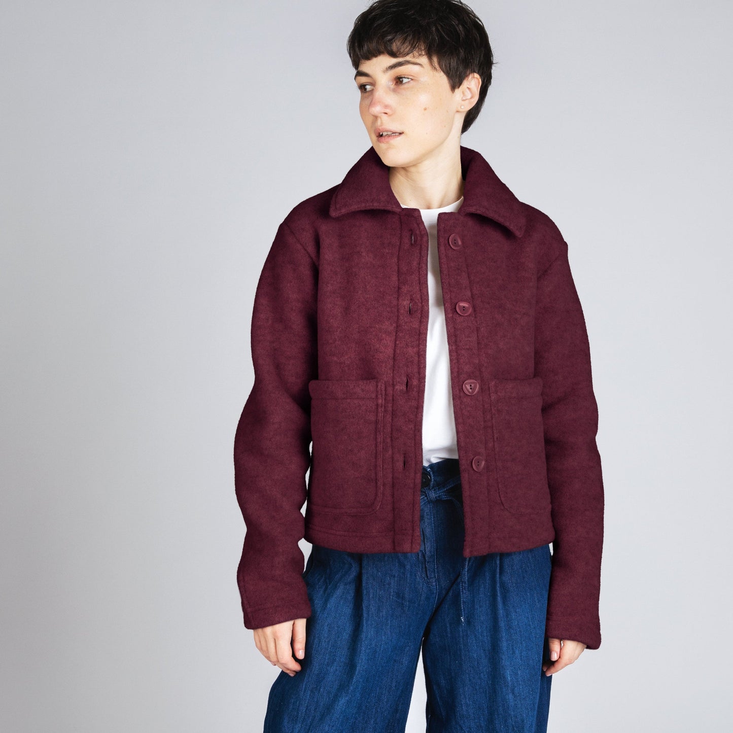 A model stands against a light gray studio background wearing a cropped maroon corduroy jacket over a white shirt, paired with wide-leg dark blue jeans and off-white sneakers.