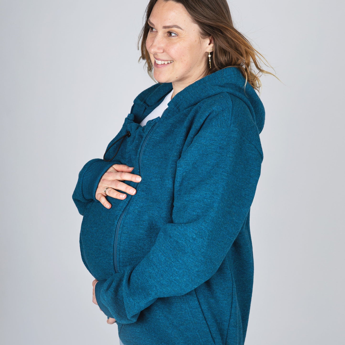 A pregnant woman wearing a teal blue heathered zip-up hoodie with a built-in belly support pouch, shown from a side-back angle against a light gray studio background.