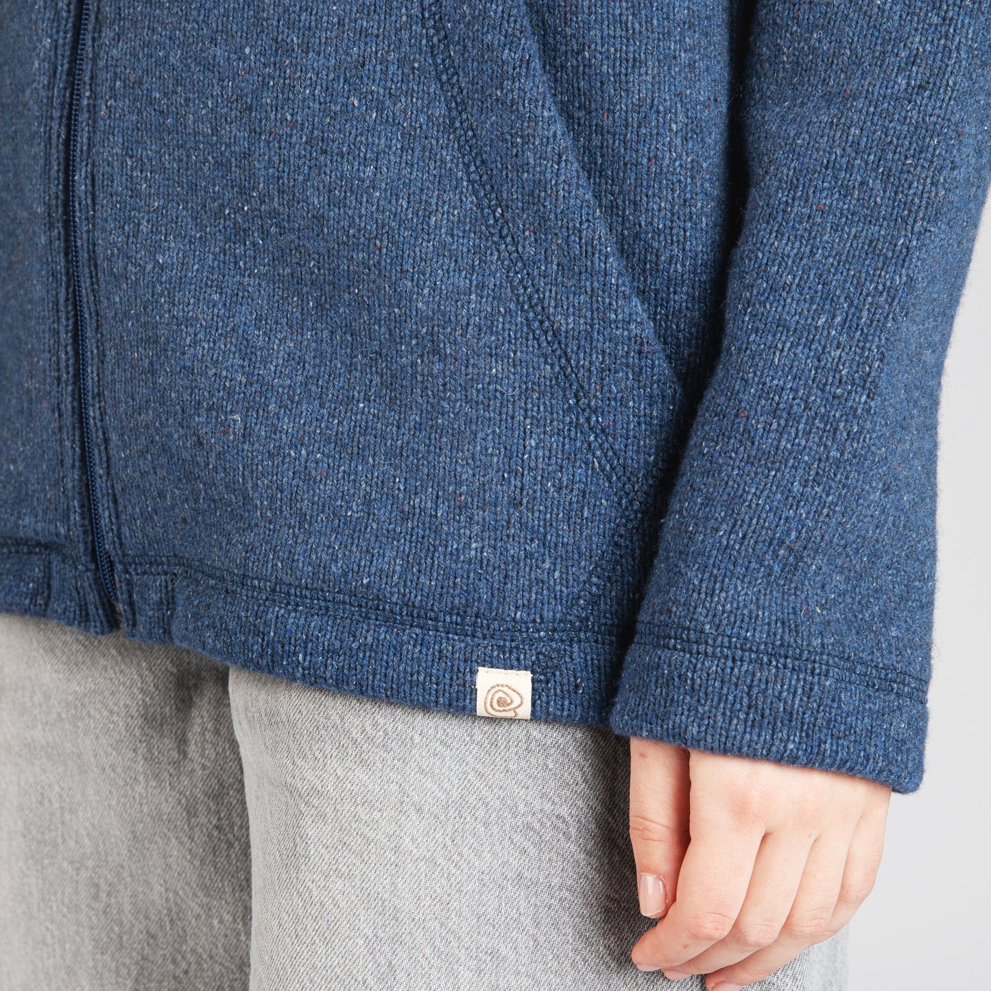Close-up of a person wearing a heathered blue zip-front jacket with a small square brand tab near the hem, paired with light gray pants, against a plain light gray background.