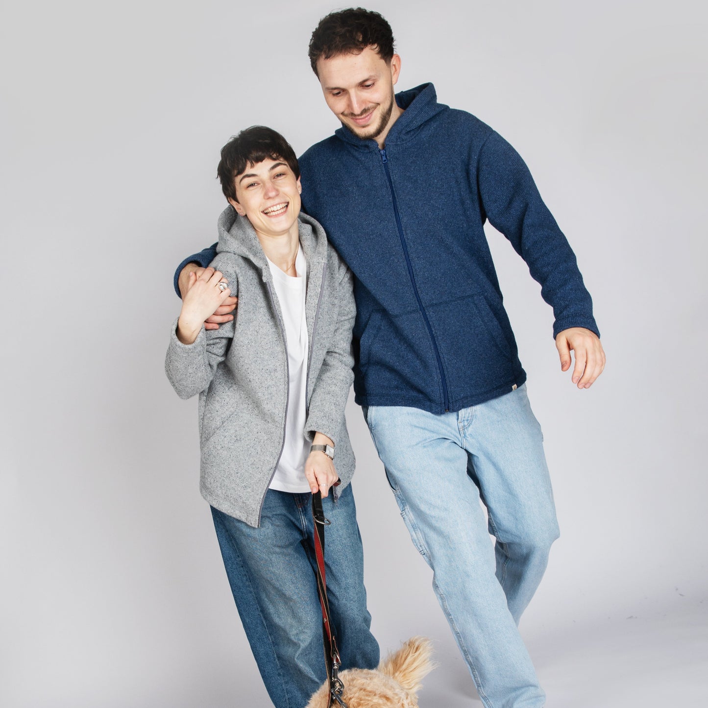 Two people walking arm-in-arm with a fluffy light brown dog on a leash against a plain light gray studio background.