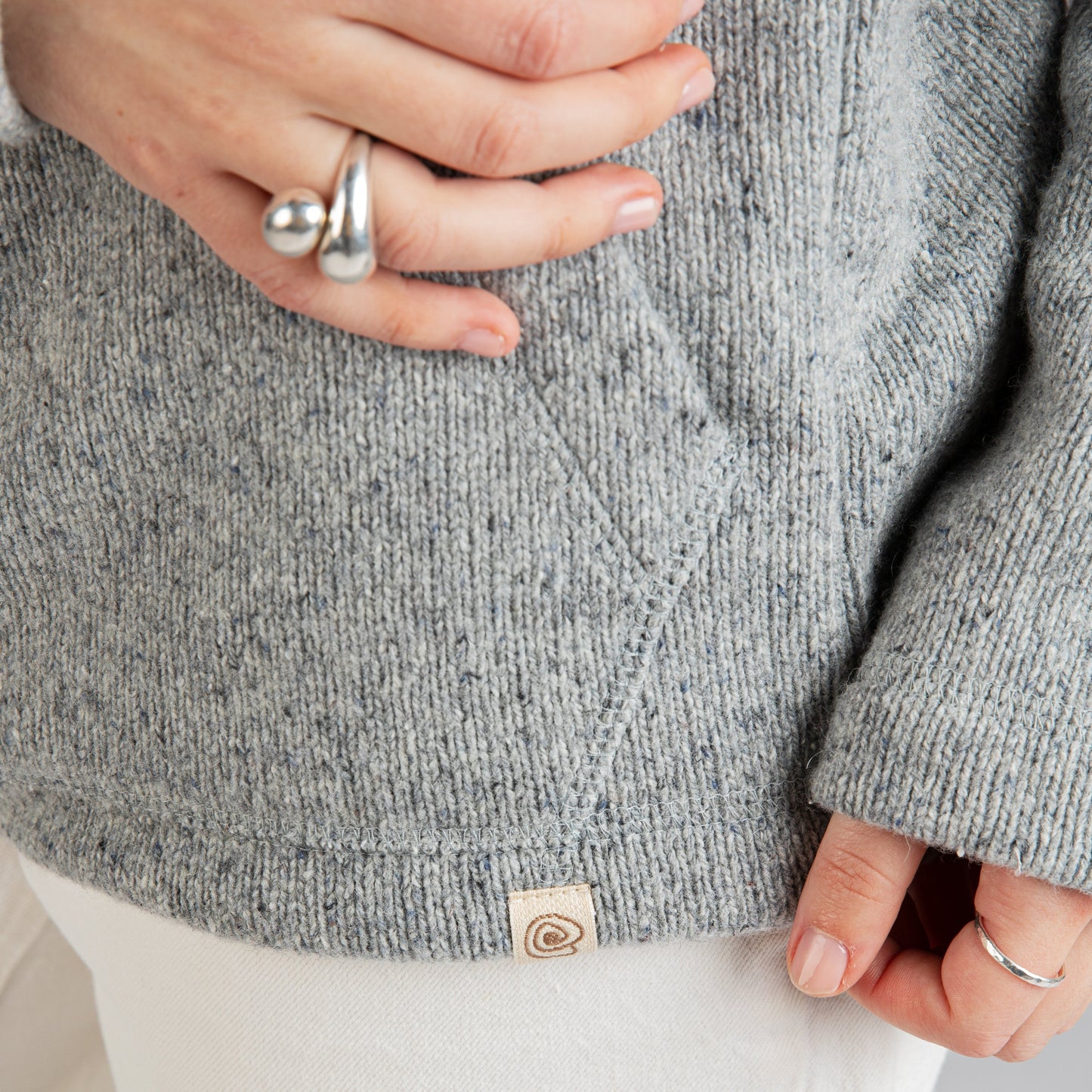 Close-up of a person wearing a heather gray knit sweater and white pants, with hands visible and a small circular fabric tag near the hem.