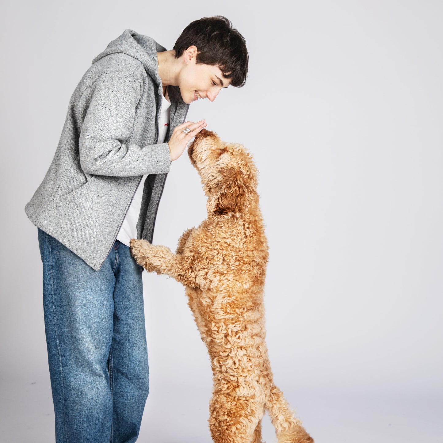 A young person in a gray hoodie and blue jeans leans down to greet a golden retriever dog standing on its hind legs and placing its paws on his hand, set against a plain light gray studio background.