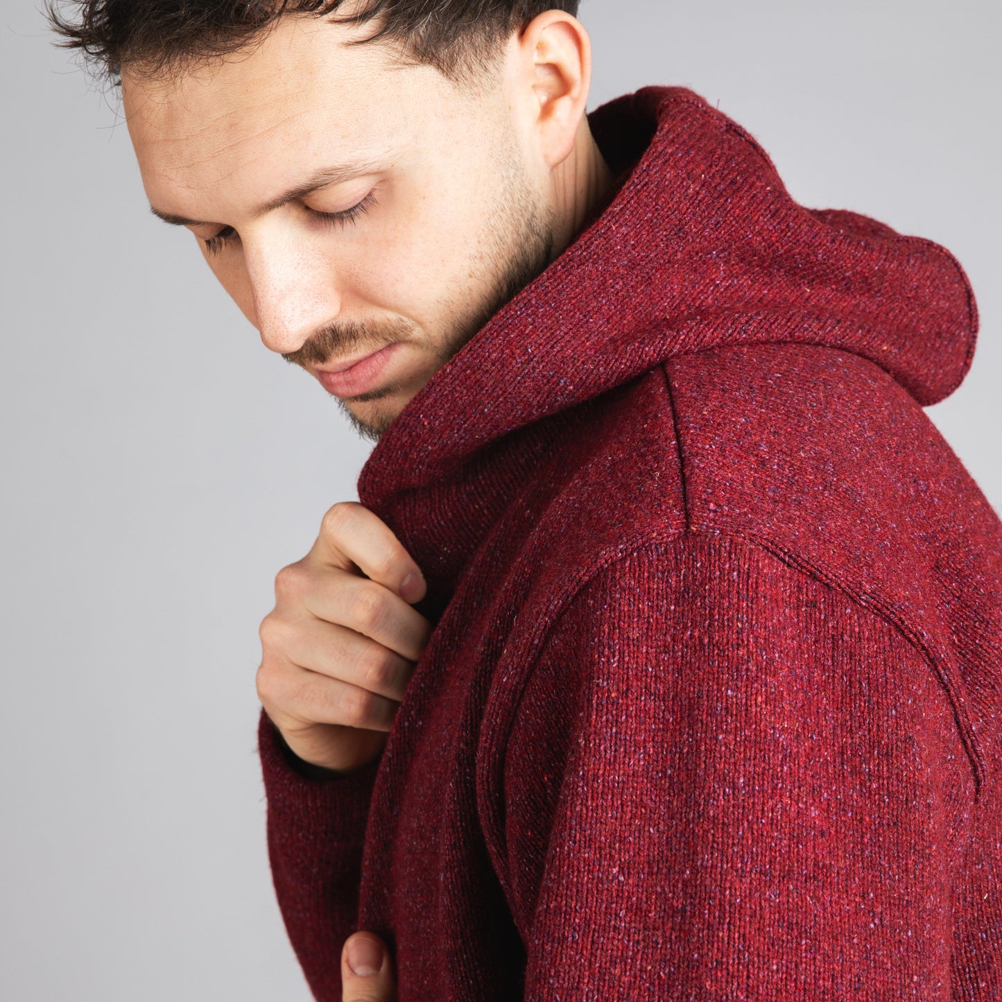 A man wearing a textured, heathered burgundy knit hoodie, looking down and adjusting the collar with one hand against a plain gray studio background.