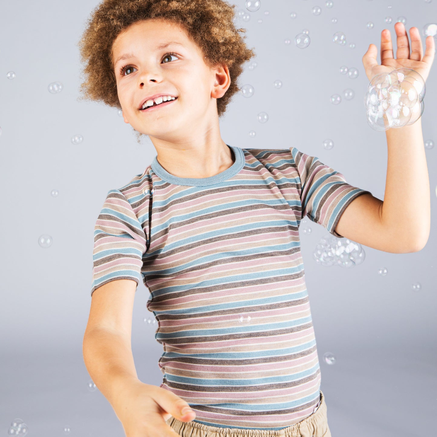 A young boy with curly brown hair smiles and looks up while playing with floating bubbles against a light gray background. He is wearing a short-sleeve striped t-shirt in muted blue, brown, and beige tones, paired with khaki pants.