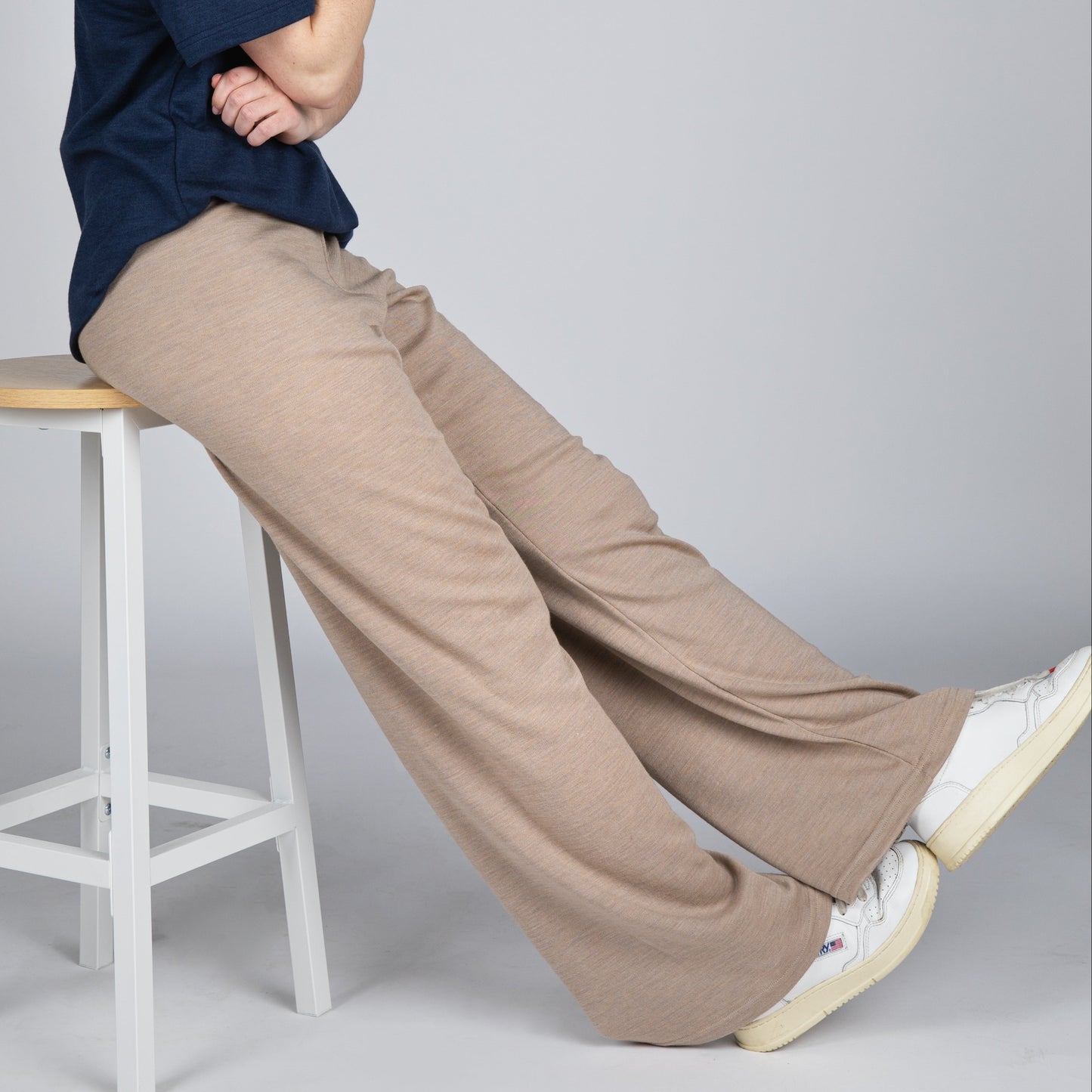 A model with short dark hair sits on a white wooden stool, wearing a navy blue short-sleeve V-neck top, beige wide-leg trousers, and white sneakers against a solid gray studio background.