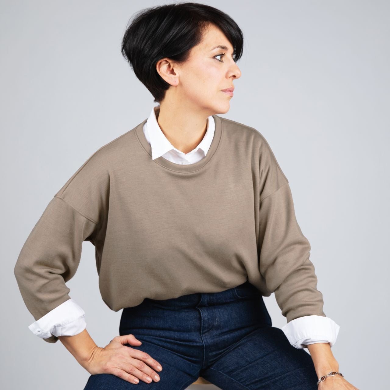 A woman with short dark hair sits on a white stool against a light gray studio background, wearing a taupe crewneck sweater over a white collared shirt and dark blue jeans.