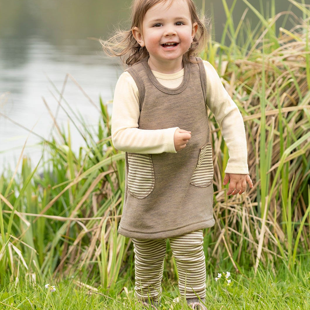 A toddler girl wearing a brown sleeveless pinafore dress over a cream long-sleeve shirt and striped leggings, standing outdoors in a grassy area next to a lake.